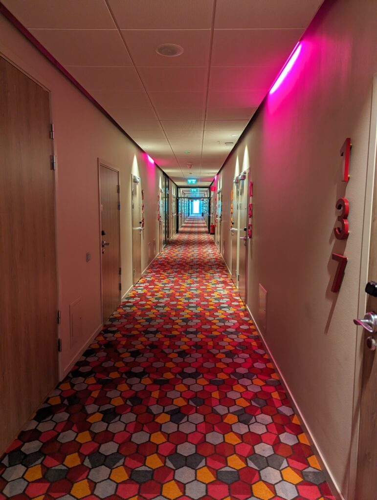 Ein Hotelfluf mit rosa Beleuchtung, einem Teppich mit Sechseckmuster. Die Sechsecke sind in rot, orange und verschiedenen Grautönen gefärbt, auf jeder Seite mindestens 25 Zimmertüren