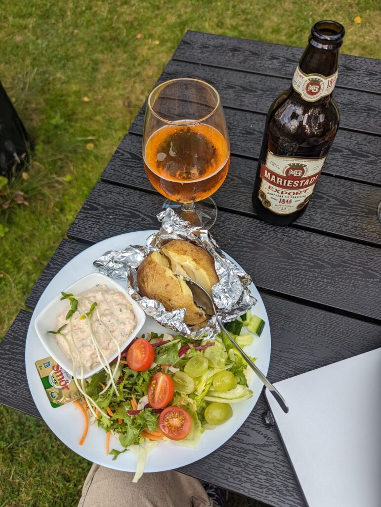 Abendessen in Silleruds Station, ein Teller mit einer Baked Potatoe, Salat mit Tomaten und Trauben, einer kleinen Schüssel mit Thunfischsalat sowie einer Bierflasche und einem gefüllten Glas Bier.