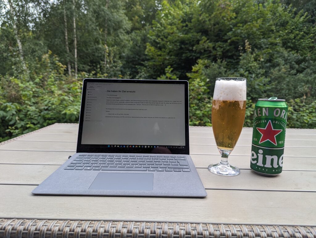 Ein Laptop aufgeklappt auf einem Tisch, auf dem Bildschirm ist der Text des Blogs zu erkennen in einem Markdown-Programm, daneben ein Glas mit Bier sowie eine Bierdose, dahinter Bäume und Sträucher.