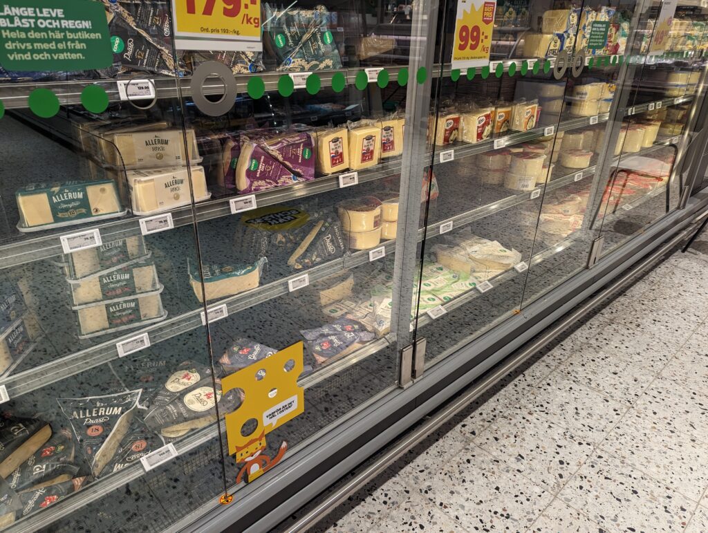 Das Käseregal in einem Supermarkt. Hinter Glastüren sind verschiedene Käselaibe zu erkennen, man sieht, dass es jeweils größere Blöcke sind.