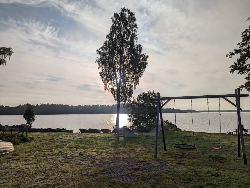 Blick in die Sonne hinter einem Baum. Im Vordergrund eine Schaukel, dahinter der See. Am gegenüberliegenden Ufer erkennt man die Bäume