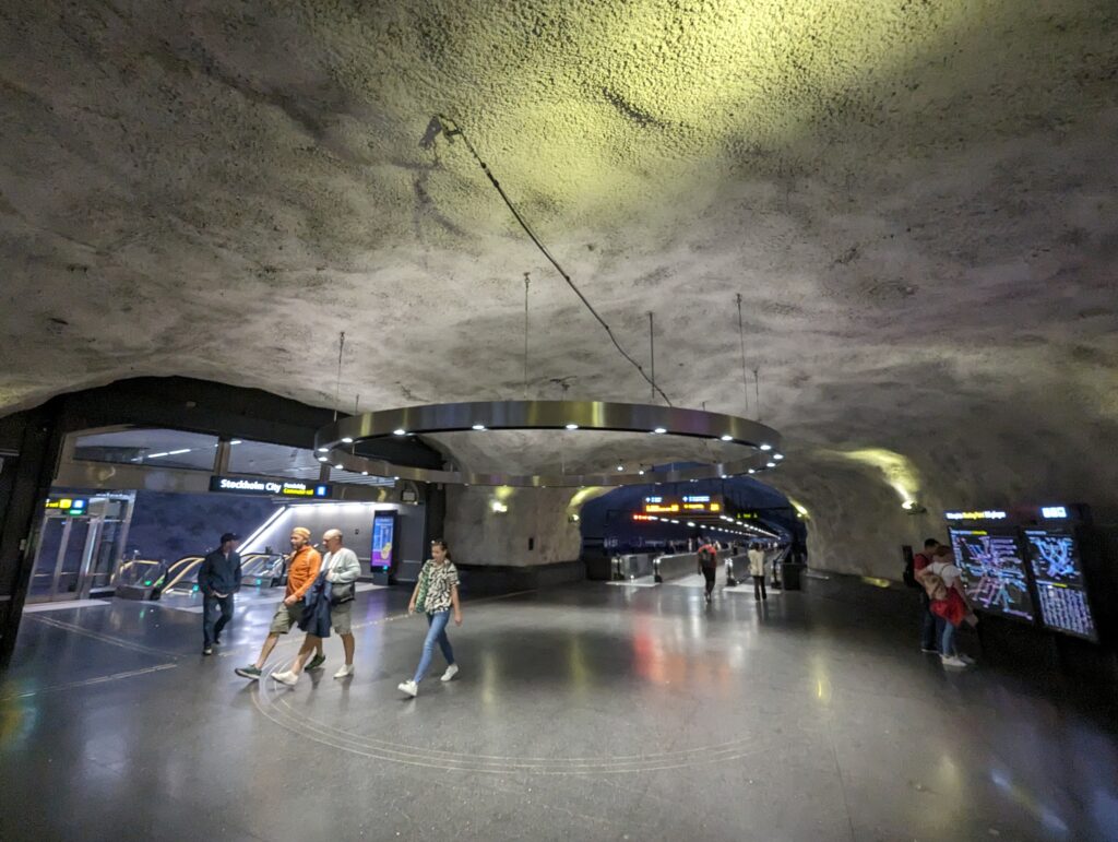 Eine grüngelb angestrahlte Decke in einer U-Bahnstation, grober Beton, darunter hängt eine rund Lampe, wie eine Felge, aus Edelstahl, Durchmesser ca. 5 Meter.