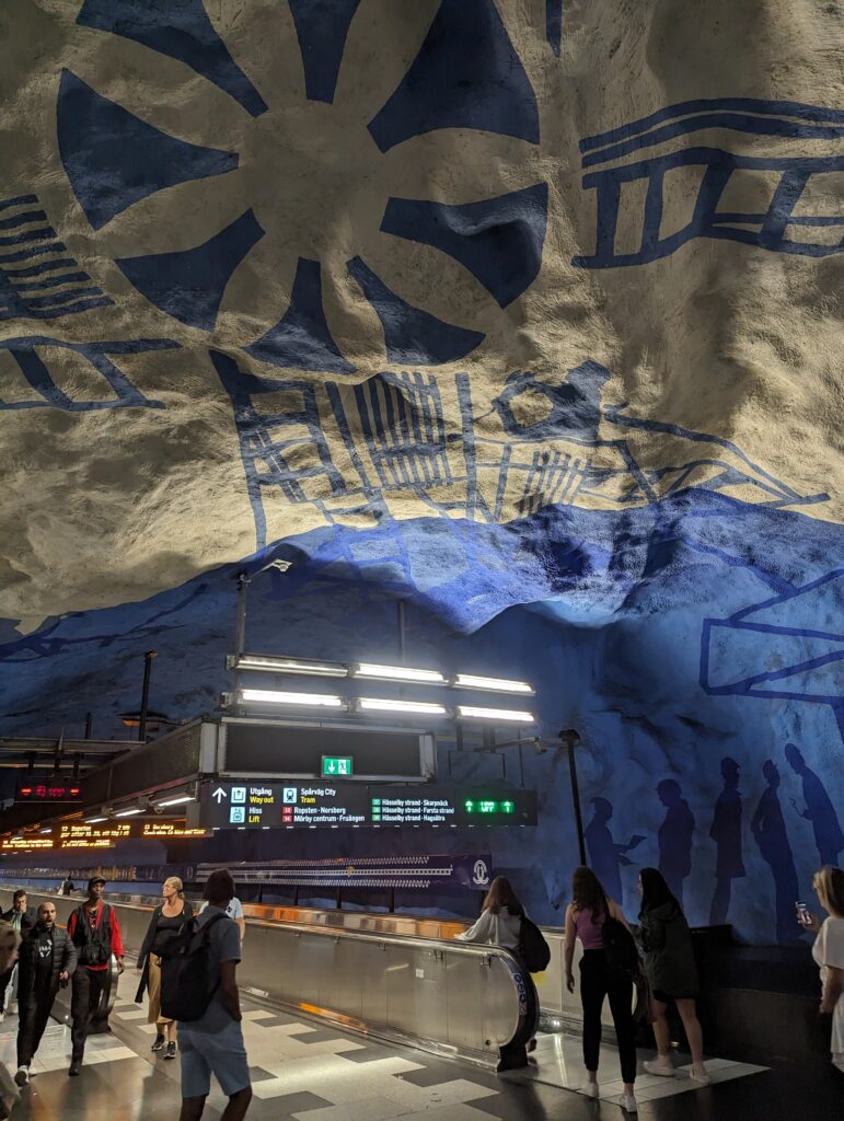 U-Bahn-Station Central mit einer Decke, die aussieht wie unebenes Gestein mit blauen Bemalungen auf weißem Grund. Eine Sonne und abstrakte Striche