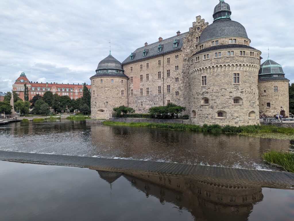 Das Schloss von Örebro umflossen von Wasser, drei Dicke Türme sind zu sehen, dazwischen der 4-stöckige Bau und das Dach. Im Wasser vor dem Schloss eine Stufe, über die das Wasser fließt.