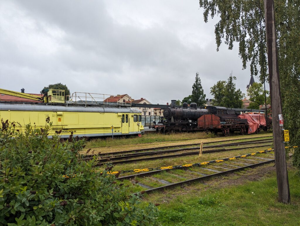 Blick auf Gleise, links ein gelber Werkstattwagen aus einem ehemaligen Schienenbus gebaut, rechts ein alter Güterwagen mit offener Ladefläche, dazwischen eine Dampflok