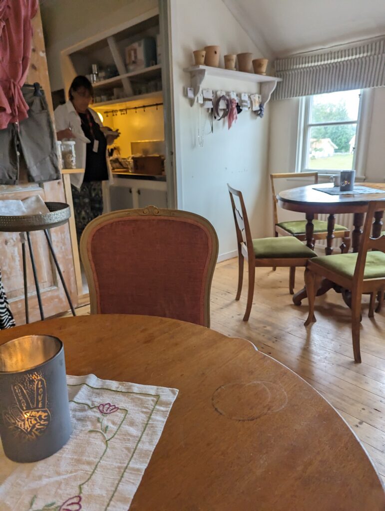 Ein Frühstücksraum. Im Vordergrund ein Tisch und ein Stuhl daran, der Tisch ist aus altem Holz, der Stuhl mit rotem Samt bespannt, dahinter ein weiterer alter Tisch mit Stühlen, links hinten der Blick in eine alte Küche.