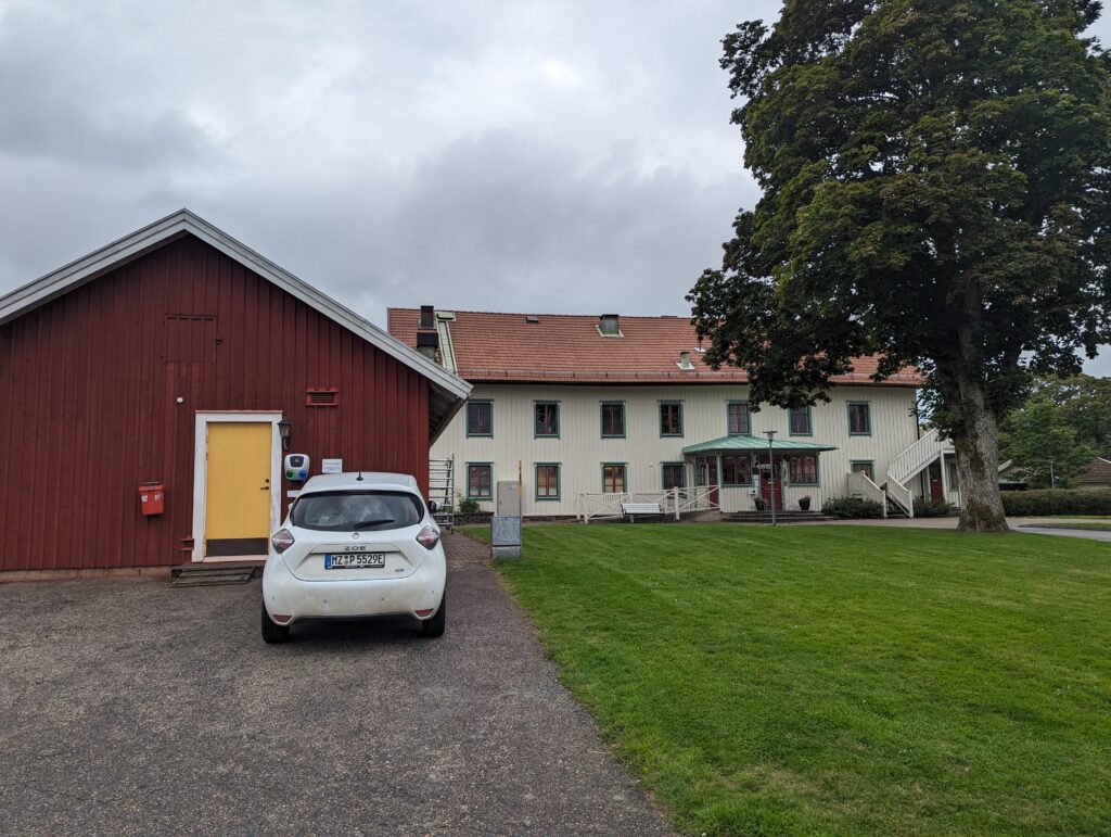Die Zoe hängt an einer Ladestation, die an einem Schuppen im typischen Schwedenrot befestigt ist, eine gelbe Tür in der Scheune. rechts daneben im Hintergrund ein großes zweistöckiges Haus, davor ein großer Baum.