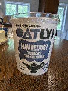 Ein größerer Joghurtbecher mit Plexiglasdeckel auf einem Holztisch. Aufschrift: The original OATLY! Havregurt Turkisk/Turkkilainen 10% fett/ravsaa 400g wow no cow!