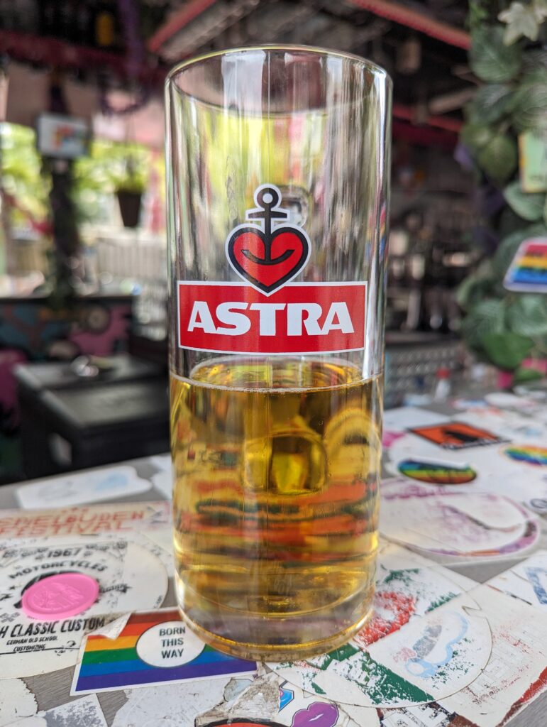 Ein Glas mit der Aufschrift ASTRA, etwa zur Hälfte gefüllt mit Bier auf einer Theke.