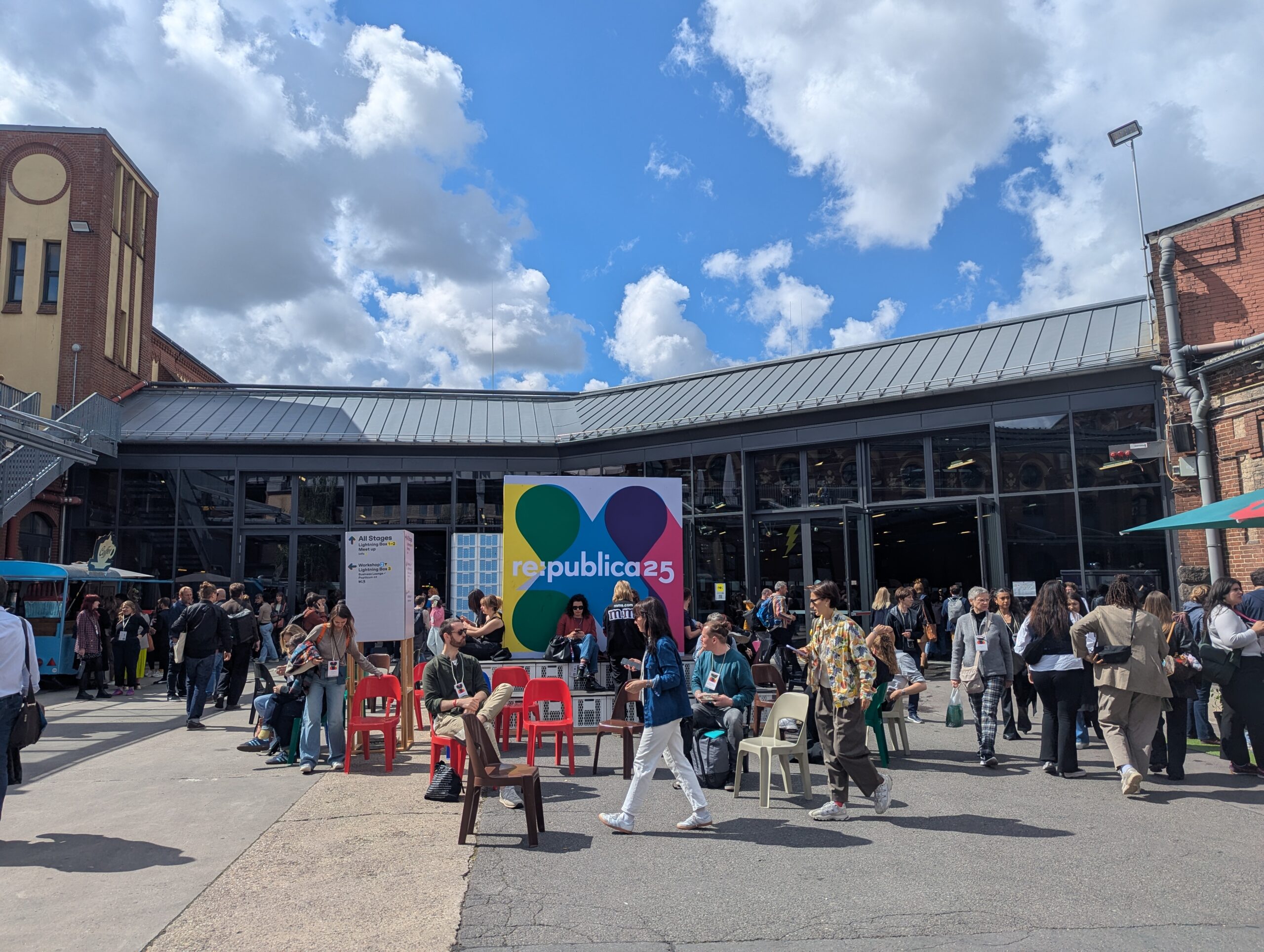 Eingangsbereich einer Veranstaltungshallte, eine eineinhalbstöckige Glasfront mit einem Blechdach, davor Menschen auf einem freien Platz, darin ein großes Plakat mit der re:publica25-Aufschrift und dem bunten XYZ-Logo